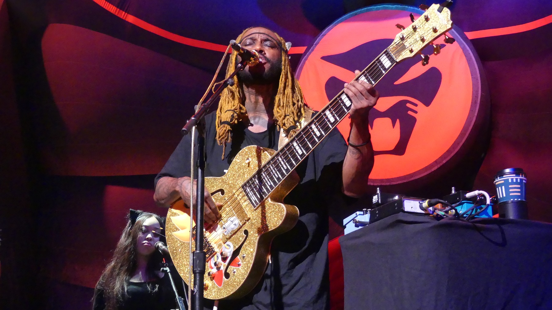 Thundercat sings and plays a gold bass against the red cat backdrop.
