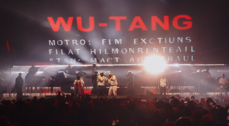 Wu-Tang Clan performing on a stage lit in red and white, with a crowd in the foreground.