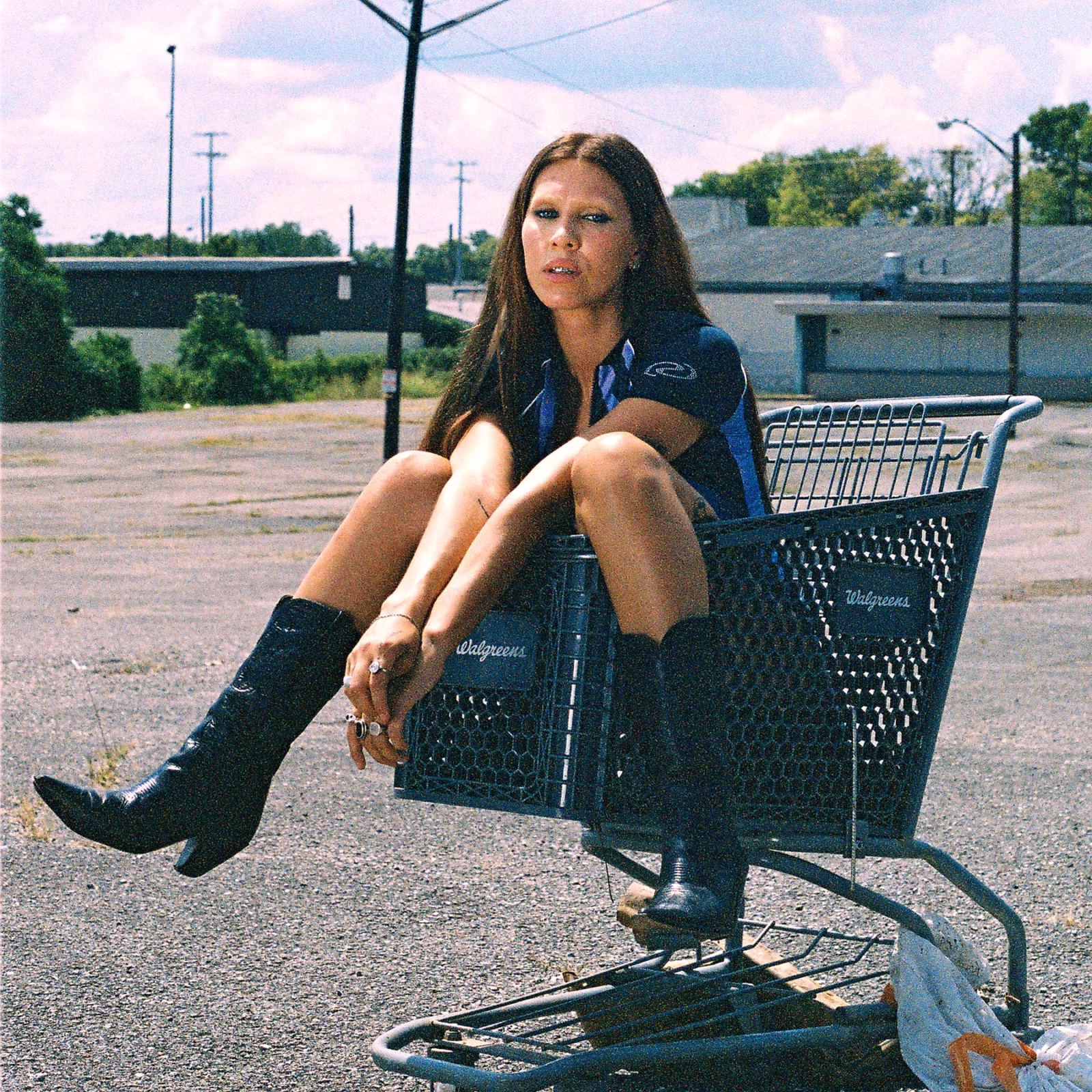 BRÍET in cowboy boots, seated in a shopping cart in an outdoor parking lot.