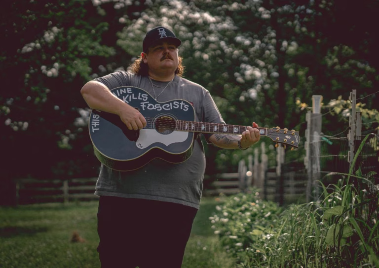 Bryan Andrews standing outdoors with an acoustic guitar.