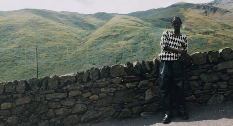 Fashion Britain campaign image: a model in a diamond-patterned knit beside a stone wall in a green hillside landscape.