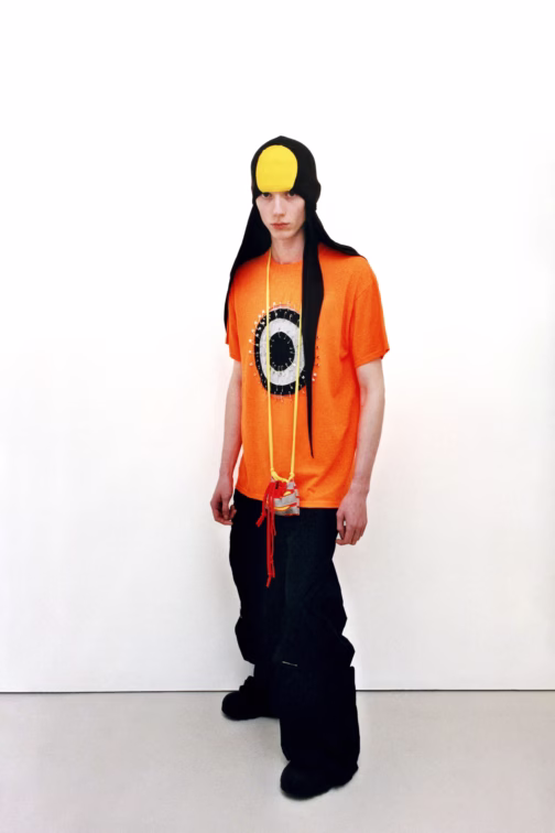 Model wearing a bright orange Jawara Alleyne top with black trousers and sculptural headwear