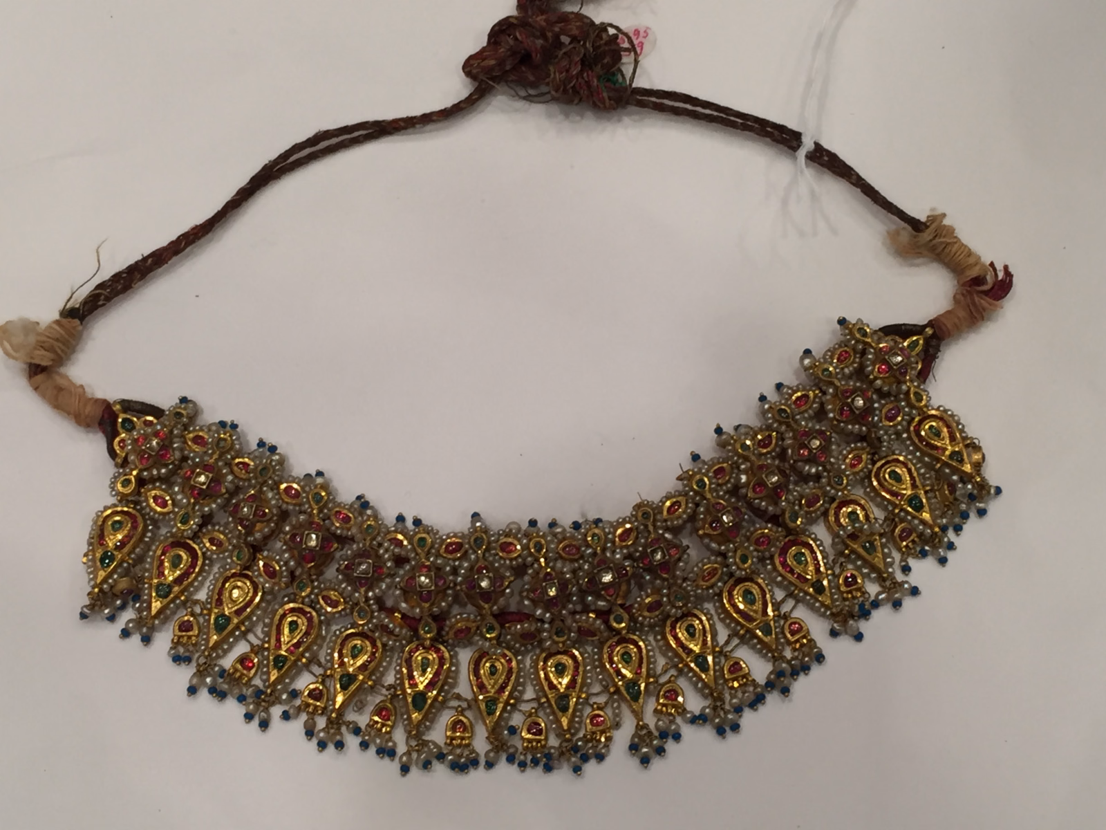 Curving gold necklace with repeated jeweled elements in red, green and pale colors, small blue drops along its edge and a textile tie.