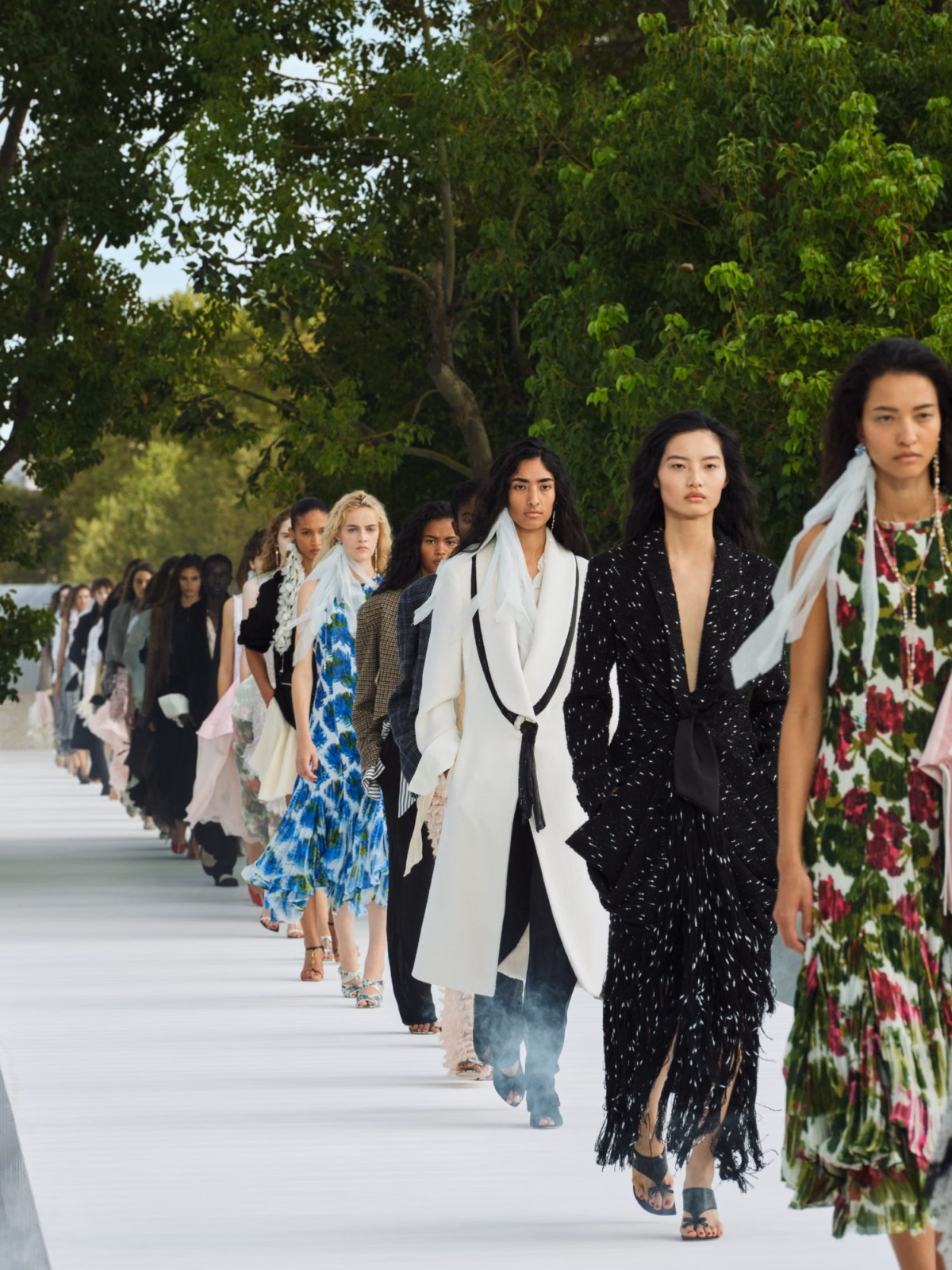 Models in floral dresses and contrasting black and white tailoring walk Dior’s tree-lined Summer 2027 runway.