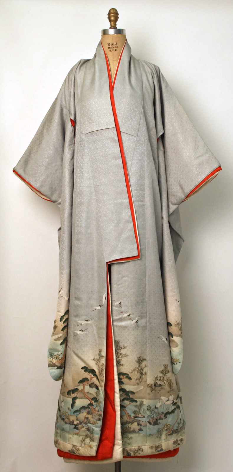 Pale silk kimono on a dress form, with red edging and lining and a landscape with trees and birds around the lower part.