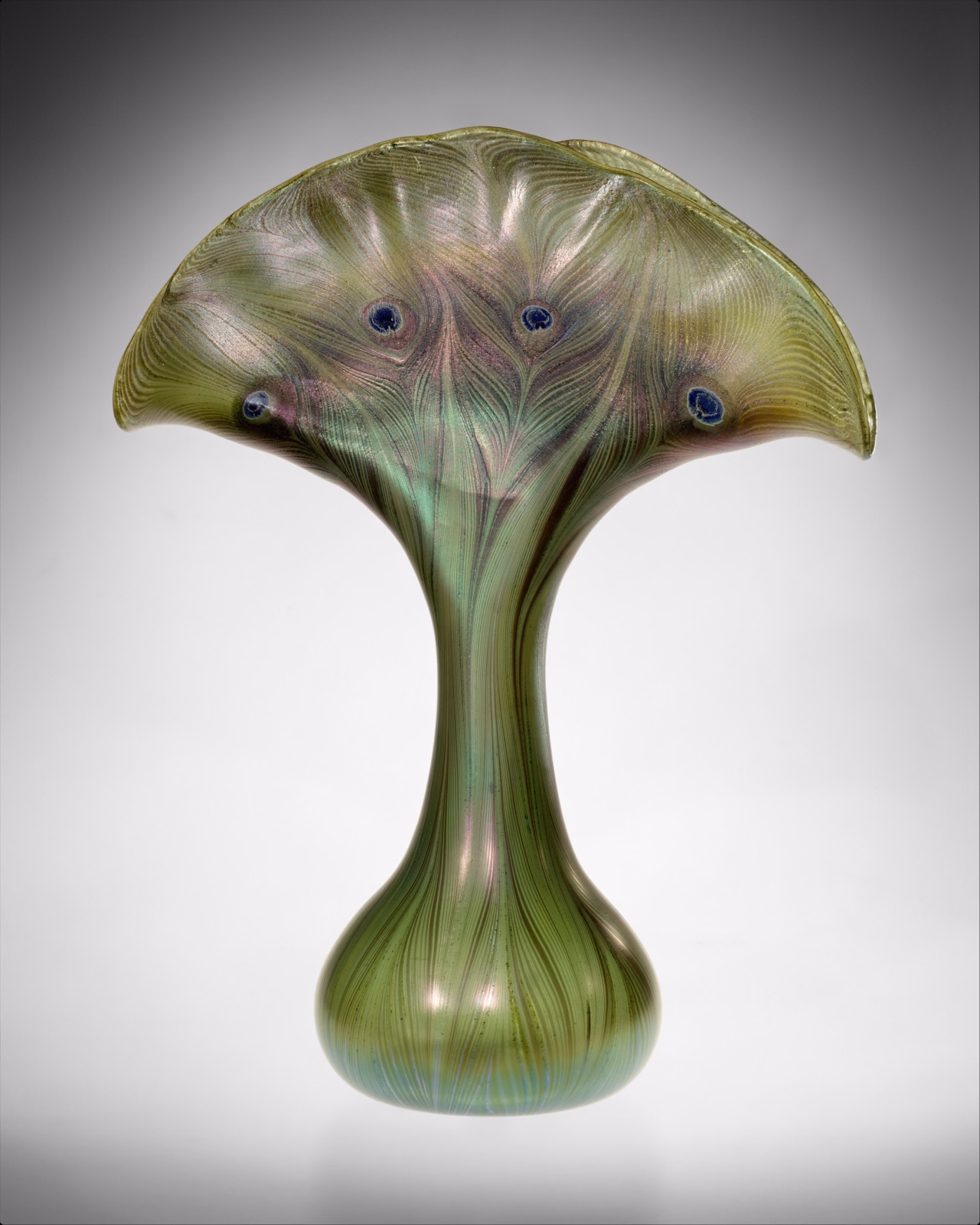 Green iridescent Tiffany glass vase with a narrow stem, rounded base and broad fan-shaped rim decorated with dark eye-like spots.