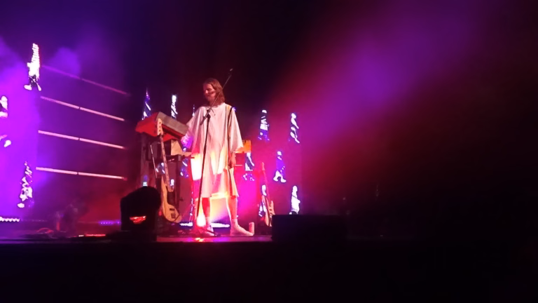 Daði Freyr smiles at the microphone beside his synthesizers, surrounded by magenta stage lighting.