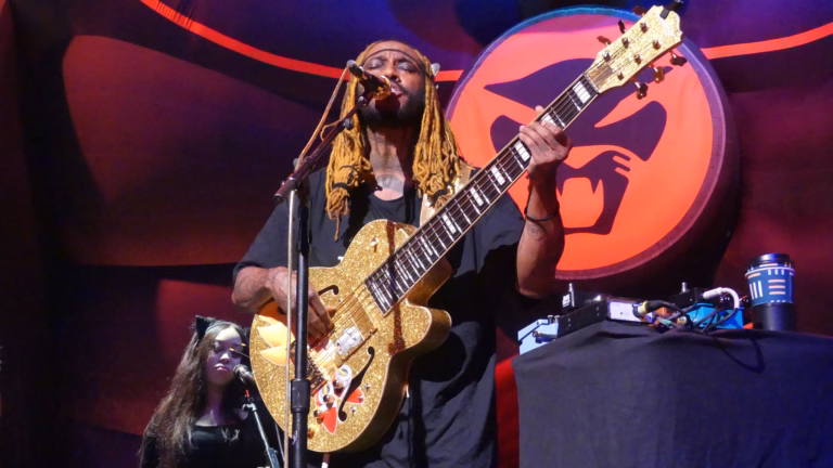 Thundercat sings into a microphone while playing a gold bass in front of a red cat emblem.