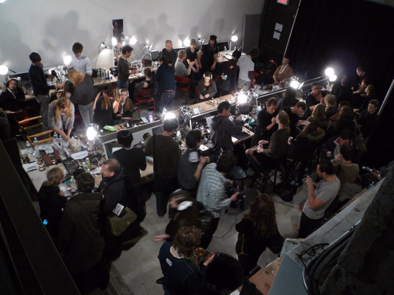 An elevated view of models and hair and makeup teams working around illuminated mirrors backstage at the Philosophy show during New York Fashion Week in 2008.
